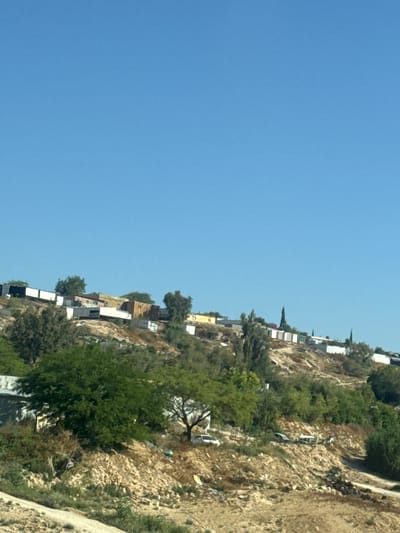 The image depicts a hillside in what appears to be Al-Qasoum Regional Council, Israel. The hillside is sparsely populated with residential buildings built along its slope. Vegetation covers the ground, with clusters of trees and shrubs interspersed among the rocky terrain. The sky is clear and blue.