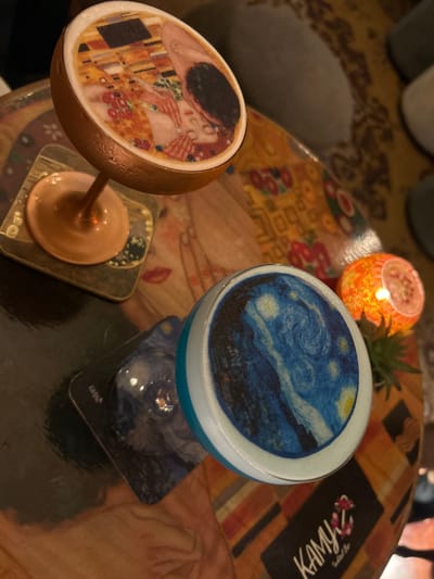 Two cocktails are displayed on a patterned table. The cocktail on the left is in a copper-colored glass and features an edible image of "The Kiss" by Gustav Klimt. The cocktail on the right is in a clear glass and has an edible image of Vincent van Gogh's "The Starry Night." A small, lit orange candle holder is visible in the background, casting a warm glow. A succulent plant is positioned behind the candle. A black coaster with a floral design and the text "KAMY" is visible under the right-hand cocktail. The table has a warm, patterned surface with a mix of yellow, orange, and brown tones, suggesting an indoor setting, possibly a bar or restaurant. The lighting is dim, with warm tones from the candle and ambient light. The overall atmosphere appears relaxed and sophisticated, evoking an artistic or cultural theme. The scene is likely set in Valletta, Malta, given the context.