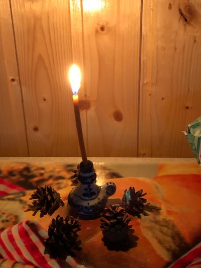 A lit candle stands in a decorative ceramic holder, shaped like a bird. The holder is blue and white with floral patterns. The candle is thin and brown, with a bright yellow and orange flame. Several pinecones are scattered around the base of the holder. A red and white striped ribbon is partially visible in the foreground. The background consists of a wooden wall with vertical planks and visible knots. The surface on which the objects are placed is covered with a brightly colored fabric, possibly a tablecloth, with abstract orange and yellow patterns. The lighting is warm and atmospheric, likely from the candle and possibly another light source from above and to the left. This scene appears to be set indoors. The location in the city of Staroe Istomino, Russia is not ascertainable from the visual information presented in the image.