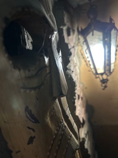 The image depicts a close-up, low-angle view of an ornate, octagonal lantern with a warm light source inside, suspended from a ceiling. The lantern has dark, possibly wrought iron, framework and glass panels. To the left, a heavily textured, possibly carved wooden surface is prominent in the foreground, with a dark opening or recess. This wooden element has a rough, weathered appearance and is decorated with what appear to be small, spherical studs arranged in a pattern. The background is a textured wall with a yellowish-brown hue, further illuminated by the lantern's light. The overall impression is one of an interior space with architectural details, possibly historical or decorative, located in Slov'yanoserbs'k, Ukraine. There are no people or overt actions visible.
