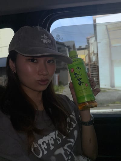 A woman in a grey "Stussy" hat holds a green "Oi Ocha" beverage bottle. She wears a grey shirt with white designs. The setting seems to be inside a vehicle, as suggested by a window reflecting an outdoor view of a building and sky. The image is slightly rotated, presenting a somewhat sideways perspective. The overall tone is relaxed and casual.