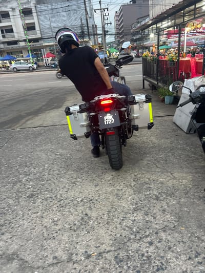 A person wearing a black t-shirt and a helmet is sitting on a motorcycle with its rear facing the camera. The motorcycle has two silver saddlebags with yellow reflective strips on the sides, and a license plate that reads "PRK 1727". The motorcycle is on a paved road next to a sidewalk with a row of buildings in the background. The buildings appear to be shops or restaurants with outdoor seating. The sky is overcast, suggesting it is either early morning or late afternoon. The scene is located in Ban Thai–Changlon, Thailand.