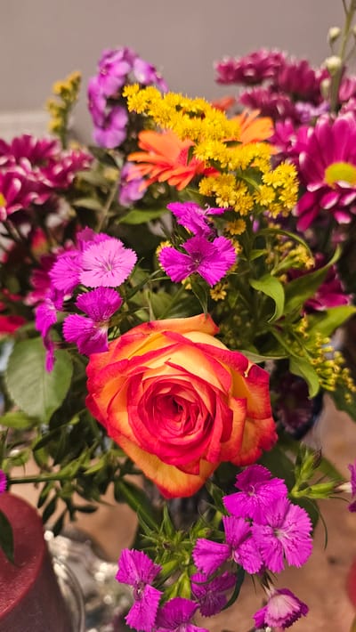 This is a close-up, eye-level, vertical, medium shot of a vibrant bouquet of flowers. The primary focus is a large, bi-colored rose in the foreground, with petals transitioning from deep red at the edges to a bright yellow-orange center. Surrounding the rose are smaller, bright pink, carnation-like flowers with serrated edges, and clusters of small yellow flowers. In the background, there are blurred purple and magenta chrysanthemums and orange gerbera daisies. The lighting is soft, creating a warm and inviting atmosphere. The background is a muted, neutral color, likely an indoor wall. There is no indication of the city of Charleroi, Belgium in the image.