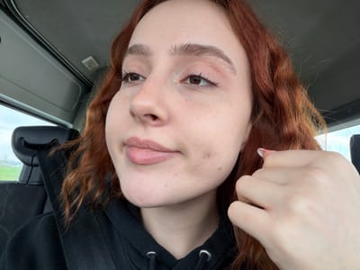 A young woman with auburn, wavy hair and light skin poses for a selfie in a vehicle. She wears a black hooded sweatshirt. Her left hand is raised near her face. The interior of the car is visible, with a rearview mirror and a gray ceiling or dashboard. The image is slightly angled.