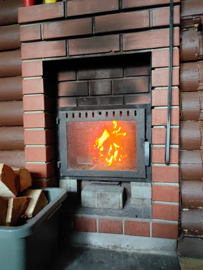 A burning fireplace is the central focus of this image, set within a rustic, log-cabin style interior. The fireplace is constructed of red brick, with a metal door featuring ventilation slits above the glass front, through which flames are visible. A black metal poker rests vertically against the right side of the brickwork, attached to the fireplace structure. To the left of the fireplace, a grey plastic bin contains several pieces of cut firewood. The surrounding walls are made of dark brown, rounded logs. The overall impression is one of warmth and coziness, likely in a home or a rural setting. The image does not contain any people or any specific indicators of a city location, beyond the requested mention of Фаніпальскі сельскі Савет, Belarus.