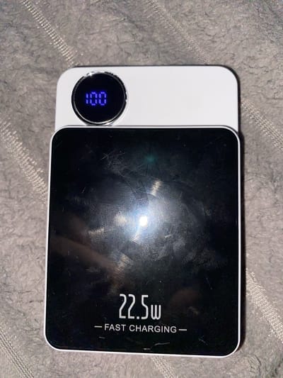 The image shows a black and white power bank with a digital display showing "100". The power bank has "22.5w - FAST CHARGING -" printed on its surface. The power bank is placed on a grey, textured surface, possibly a blanket or carpet. There is a bright light reflection on the black surface of the power bank. No specific location in Fălești, Moldova can be determined from the image.