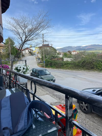 The image presents a daytime view from a balcony or elevated position overlooking a rural or semi-urban street in Djigurovo, Bulgaria. The sky is clear and blue with scattered wispy clouds, suggesting fair weather.
In the immediate foreground, a dark metal railing, possibly wrought iron, frames the bottom and right side of the shot. Resting on a glass table behind the railing is a blue and dark blue backpack. Below the railing, a colorful red, yellow, and green children's ride-on toy car is visible on the ground or a lower level. White artificial flowers are arranged in a pot on the railing, and some purple flowers are also seen below.
The midground features a narrow, paved road that curves slightly to the left. Several cars are parked along or near the road: a dark green sedan is parked on the asphalt, with part of its license plate visible showing Cyrillic characters including "P" and numbers that appear to be "478 BC". To its right, a black car is partially visible, parked on a gravel shoulder. A tall, bare deciduous tree stands prominently on the left side of the street, suggesting late winter or early spring. Utility poles and power lines crisscross the sky. Houses with tiled roofs and surrounding vegetation line both sides of the street.
In the background, lush green hills and mountains rise, with some higher peaks displaying lighter patches, possibly snow or bare rock. The overall setting is a quiet, residential area with natural landscapes in the distance. No specific activity or event is taking place, depicting a static street scene. A partial text "sysmo" is faintly visible on a cylindrical object in the lower left.