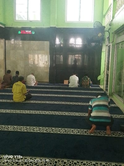 A wide-angle interior photograph captures a prayer room or mosque in Jagapura Kidul, Indonesia. Six male individuals are observed from behind, seated or kneeling on a dark blue prayer carpet with repetitive white-patterned horizontal stripes. All individuals face away from the camera, suggesting engagement in prayer or quiet contemplation.
The upper walls are painted light green, while the lower section features dark, reflective paneling on the right and a lighter tiled section on the left. Two large, rectangular windows are set high on the upper wall. A digital display with green Arabic script and numerals, possibly indicating prayer times or other information, is mounted on the dark paneling. A black wall-mounted fan is positioned in the upper right corner. A glass-paned door or partition is visible along the right wall.
In the foreground, one individual wears a yellow short-sleeved shirt with text and dark pants, seated with hands in his lap. Closer to the camera and slightly to the right, another individual wears a horizontal striped shirt (teal, grey, white) and dark pants, kneeling. In the mid-ground, four additional individuals are seated; one in a white shirt, one in a green and brown patterned shirt, and two partially obscured figures to the far left. All individuals appear to be barefoot. The scene is illuminated, with natural light entering from the windows. The image is watermarked "vivo Y19s Akhter & Arvan 30 Des 2025, 15:45".