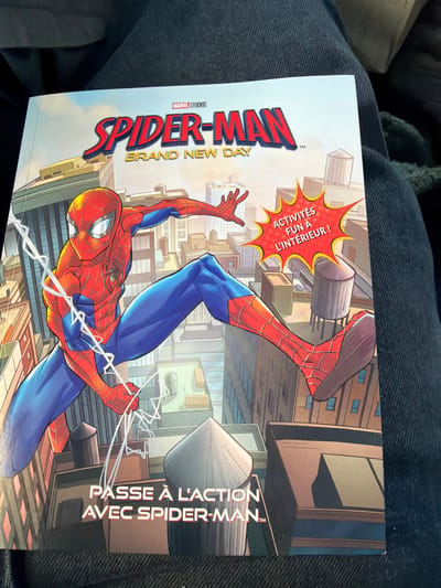 The image shows a close-up of the cover of a "Spider-Man: Brand New Day" activity book. Spider-Man is depicted mid-air, leaping between buildings in a stylized city. The cover is in French, with the title "SPIDER-MAN BRAND NEW DAY" prominently displayed. Below the main title, it says "ACTIVITÉS FUN À L'INTÉRIEUR !" (Fun activities inside!). At the bottom of the cover, there is more French text: "PASSE À L'ACTION AVEC SPIDER-MAN" (Get into action with Spider-Man). The book is lying on dark, textured fabric, possibly clothing. The city depicted is a generic urban environment with skyscrapers and water towers, and there is no specific information linking it to Ödsmål, Sweden.