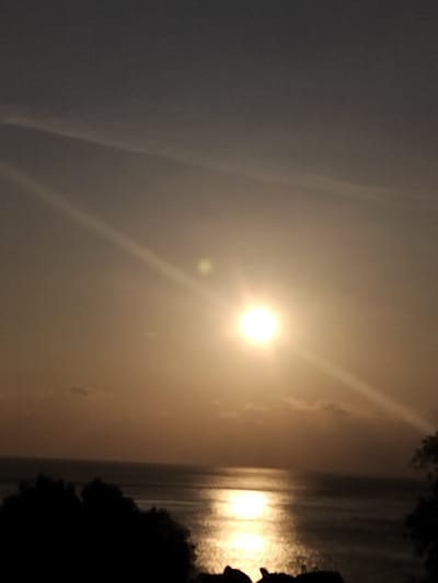 The image displays a scene featuring a prominent, bright circular sun centered in the upper mid-frame, suggesting either sunrise or sunset over a body of water, observed from Pyli, Greece. The sky transitions from a dark gray at the top to a golden-orange hue around the sun and horizon, with a few faint, wispy clouds and linear contrails visible in the upper sky. The sun's intense light creates a dazzling, shimmering golden reflection path across the calm surface of the body of water below, extending towards the viewer. The foreground presents dark, silhouetted forms of dense foliage or trees in the lower left and right corners, and irregular dark shapes, consistent with rocks or a distant landmass, along the bottom edge of the frame. No individuals, specific actions, or interactions are visible.