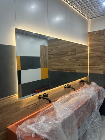The image depicts a modern bathroom under construction, likely in a commercial or public space given the multiple sinks. The scene is located in Druzhny, Russia. The primary focal point is a long, minimalist vanity with a wooden countertop and a bright orange sink basin covered in protective plastic sheeting. Two matte black faucets are mounted on the wooden countertop, each poised over an unseen drain. Above the vanity, a large, rectangular mirror reflects the interior, showing a patterned backsplash of white, black, and yellow tiles, as well as wood-effect paneling. The wall behind the mirror is clad in wood-effect tiles and dark gray panels, with a strip of warm LED lighting accentuating the ceiling line. The ceiling itself is a white grid system, typical of suspended ceilings. In the reflection of the mirror, a red and black object, possibly tool or construction material, is visible on the floor. The overall impression is one of a contemporary, stylish bathroom nearing completion, with an emphasis on natural textures and sleek design elements.