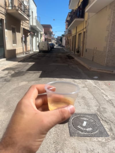 A first-person perspective shot shows a hand holding a clear plastic cup filled with a light brown beverage, possibly coffee, in the foreground. In the background, a narrow street in Stornara, Italy, is lined with buildings and features a manhole cover with textured markings. The street appears to be made of asphalt and concrete, with some debris scattered on the ground. Sunlight casts shadows across the scene, indicating it is daytime. The buildings flanking the street are of varying heights and colors, with balconies and shuttered windows visible. A white van and other vehicles are parked along the street, and a satellite dish is mounted on one of the buildings. The sky is clear and blue.
