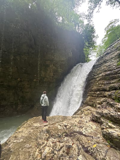 A medium, eye-level shot captures a person positioned on a rocky outcrop in front of a waterfall. The person is wearing a light-colored hooded sweatshirt, dark pants, and red shoes. They are facing the waterfall with their hands in their pockets. The waterfall is white and appears to be forceful. The surrounding environment consists of steep, rocky cliffs covered with greenery, indicative of a natural setting. Based on the location provided, this scene is likely in or near the city of Tsabal, Abkhazia.