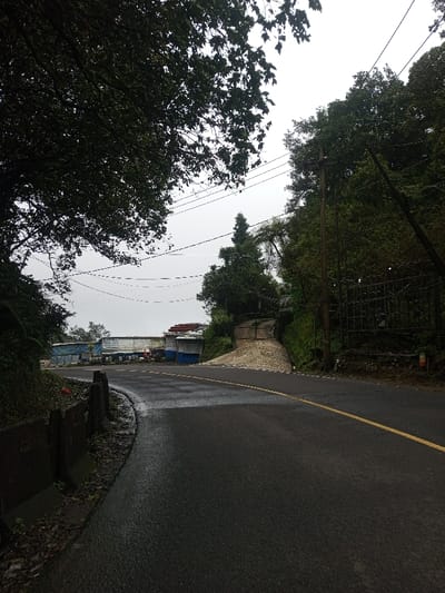 The image displays an asphalt road in Deothang, Bhutan, under an overcast sky. The road, visibly wet, features a single yellow centerline and curves gently to the left in the foreground, ascending slightly. On the left side of the road, a low concrete barrier separates the pavement from a dirt embankment covered in vegetation, beneath a large tree with dark green foliage that fills the upper left portion of the frame. Multiple utility lines are strung horizontally across the upper mid-ground. On the right side, a steep, densely treed slope rises, featuring a prominent dark utility pole and a large, dark metal gate with a patterned design, leading to an uphill, unpaved path. In the mid-ground, beyond the road's curve on the left, a collection of makeshift structures, characterized by various colored tarpaulins (blue, white, red) and corrugated metal, are visible, accompanied by several cylindrical containers on the ground. No individuals or vehicles are present.