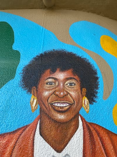 The image shows a close-up of a mural on a wall. The mural depicts a portrait of a smiling Black woman with dark, curly hair and golden earrings. She is wearing a brown garment with a white lining. The background features abstract shapes in blue, green, yellow, and beige. The location, Tawpo, Nigeria, is not discernible from the image.