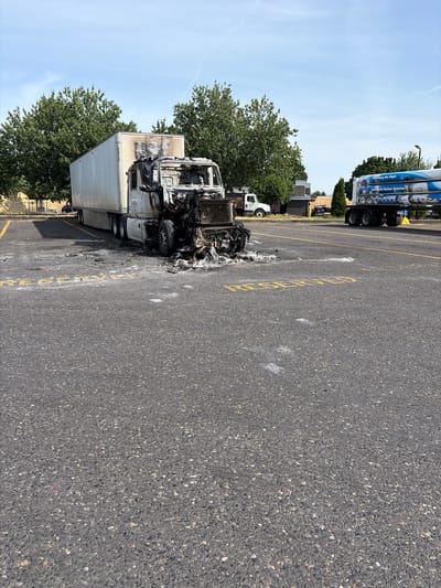 The image shows a burnt-out semi-truck and trailer parked in an asphalt lot. The truck's cab and engine area are heavily damaged, with charred metal and debris scattered around. The trailer appears to be intact but is discolored from the heat and smoke. The truck is parked between two yellow lines painted on the asphalt, suggesting it is in a parking space. In the background, there are trees, buildings, and other vehicles, including another semi-truck with large blue cylindrical tanks on its trailer. The sky is clear and blue, indicating a sunny day. The scene appears to be in a commercial or industrial area. No people are visible in the image. The time of day is likely daytime due to the bright sunlight and shadows. The overall impression is one of destruction and aftermath, likely following a fire. There is some visible text on the trailer of the intact truck, which reads "Industrial Helium Solution."