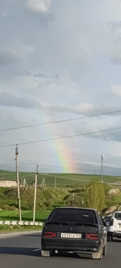 A medium-angle outdoor shot depicts a rainbow in a cloudy sky over a green hilly landscape. Power lines run across the foreground of the image. A black car with license plate E575TA 163 is partially visible in the lower foreground, along with the rear of a white car. The image appears to have been taken from a moving vehicle on a road. Based on the presence of the license plate, the location could be Azatan, Armenia, given the landscape aligns with that region.