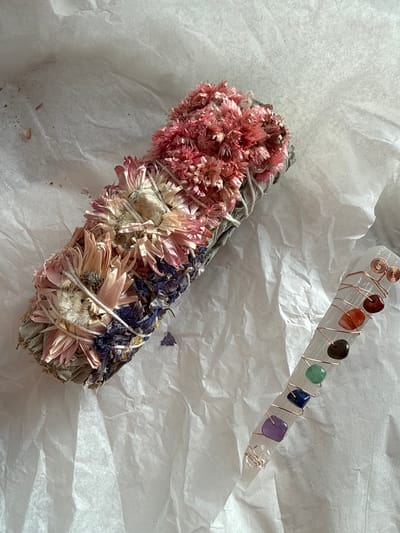 The image displays two spiritual or wellness items laid out on a crumpled, off-white, textured paper surface, likely for a product shot or display.
The primary subject in the foreground is a floral smudge stick or bundle, oriented diagonally from the middle left towards the bottom right. It appears to be made of dried sage or similar herbs, tightly bound with white string. Adorning the bundle are several clusters of dried flowers: vibrant pink, small, fluffy flowers at the top-left section, followed by larger, paler pink and cream-colored daisy-like blooms in the middle, and finally, dark purple or blue dried flowers and leaves at the bottom-right section.
In the upper right portion of the image, running diagonally towards the top right, is a slender, translucent white selenite crystal wand. This wand is intricately wrapped with thin, coppery wire that forms small spirals at one end. Along the length of the wand, embedded within the copper wrapping, are seven small, polished, square-cut gemstone beads, each a different color, representing the chakra spectrum: a reddish-orange stone, a darker brown/black stone, a green stone, a deep blue stone, a purple stone, and a clear or very pale stone at the tapered end.
The background consists solely of the crumpled paper, which shows varying shades of light and shadow, indicating bright, possibly natural lighting from an unseen source to the upper left, casting subtle shadows to the lower right of the objects. There is no visible text in the image. The arrangement suggests an aesthetic presentation of items commonly used in cleansing rituals or crystal healing practices.