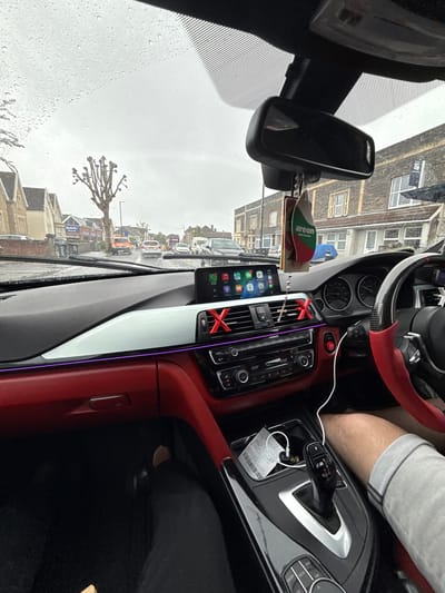 The image depicts the interior of a BMW car, viewed from the passenger seat, looking forward through a rain-splashed windshield on an overcast day. The car's dashboard features a two-tone design with dark gray and prominent red accents on the lower half, complemented by a horizontal white trim piece and an illuminated purple ambient light strip. Red "X" shaped covers are fitted over the central air vents, and an infotainment screen displays an Apple CarPlay interface with various app icons including Maps, Phone, and Music. A rearview mirror is visible, from which an "Areon" brand air freshener hangs.
The driver's right leg, clad in light gray shorts, is visible near the center console, which houses an automatic gear shifter with an "M" logo, an iDrive controller, and a storage compartment containing a receipt, keys, and charging cables. The steering wheel, partially visible, has carbon fiber elements and red accents.
Outside the vehicle, the wet road surface suggests recent rain. A moderate volume of traffic is present, including an orange van, a silver car, and a red double-decker bus further down the street. The street is lined with multi-story brick buildings, characteristic of British architecture, some appearing residential and others commercial. A large, bare, pollarded tree stands prominently in the mid-ground on the left. Business signs, including "Kwik Fit" and "McDonald's," are visible along the street.
The location is identified as **Church Road, Lawrence Hill, Bristol, United Kingdom**, looking west. The distinct architecture, the presence of a Kwik Fit garage on the left, and the specific bare tree are consistent with this area.