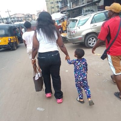 A daytime street scene in Awada, Nigeria shows four people walking away from the viewer on a paved road. A woman with long black hair wears a white top, black pants, and pink shoes, holding a brown tote bag in her left hand, and holding the right hand of a child in a long-sleeved blue and pink patterned pajama-like outfit with black sandals. To the woman's left, another woman wearing a white top and light-colored shorts walks in the same direction. To the right of the woman and child, a man wearing a red shirt, tan shorts with white stripes, and a tan baseball cap also walks in the same direction with a black bag slung across his shoulder. In the background are buildings, parked cars, a yellow tuk-tuk, and a food vendor with a white umbrella.