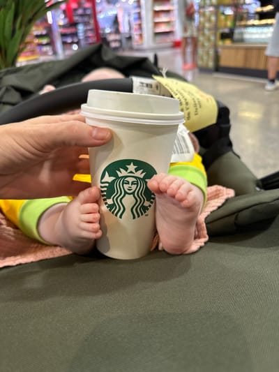 The image, taken from an overhead perspective, captures a close-up of a baby's feet resting near a Starbucks coffee cup, held by a person's hand. The baby is situated in an olive green baby carrier or stroller, with small, delicate bare feet visible. They are dressed in a bright yellow and lime green outfit, partially covered by a pink knitted blanket or fabric.

A light beige Starbucks cup, prominently featuring its green siren logo, is held in the foreground by a hand with short, neat fingernails. Behind the cup and baby, a blurred background reveals the interior of what appears to be a supermarket or retail store, with shelves stocked with various products. This suggests a shopping environment.

Near the baby's head, a yellow piece of paper with French text is partially visible, showing phrases like "Au pied," "De l'avion," and "Délivré." Another white label with a barcode is also present near the baby. The scene depicts a common everyday activity of a caregiver with an infant, likely during a shopping trip. The presence of French text suggests a region where French is spoken, which aligns with the provided context of Nouaceur, Morocco. The image creates a candid moment focusing on the contrast between the popular coffee cup and the tiny baby feet. The time of day appears to be daytime, given the indoor lighting.