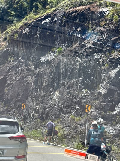 A silver SUV is parked on the right side of a paved road that curves to the left, with its rear visible. To the right of the road, a cyclist wearing a purple shirt and black shorts stands with a bicycle, facing away from the camera. Further to the right, two cyclists are standing with a bicycle. One is wearing a teal shirt and a white helmet, and the other, in the foreground, wears a dark long-sleeved shirt with "Explore" written on the back and a dark helmet. They are positioned on the road, near a steep, rocky cliff face covered in sparse vegetation. Two yellow directional signs with black arrows are visible on the cliff face, indicating a curve in the road. The scene appears to be located in Lac Duong, Vietnam, a mountainous region known for its scenic drives and cycling routes.