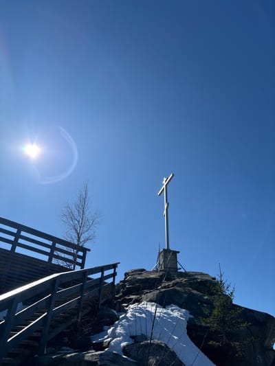 The image depicts a low-angle view of a rocky terrain on a sunny day in what appears to be the city of Gubakha, Russia. A white cross stands atop a rock face. A wooden structure, possibly a staircase or walkway, ascends the rock face. Snow is visible on the rocks, indicating a cold climate. The bright sun is in the lower part of the frame, creating a lens flare effect. The sky is clear blue.