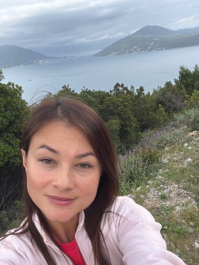 The image is a selfie of a woman with brown hair and green eyes wearing a light pink jacket over a red top. She is smiling slightly. The background reveals a scenic view of a body of water, possibly the Adriatic Sea, with a long, narrow island or peninsula in the distance. Lush green vegetation and rocky terrain surround the woman, indicating she is likely on a hill or mountain overlooking the water. Given the location, this is very likely the city of Provodina, Montenegro.