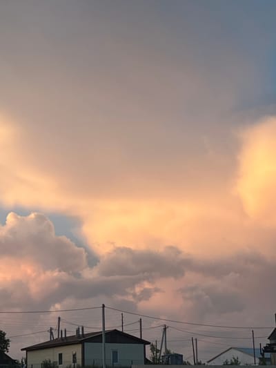 The image depicts a twilight sky over a rural settlement, likely Ludorvay, Russia. The sky is filled with dramatic, billowing clouds, tinged with soft orange and pink hues from the setting sun. Below the vibrant sky, a low-lying building with a light yellow and white facade is visible, featuring several windows. Power lines and utility poles traverse the scene horizontally, suggesting a functional, residential area. The overall atmosphere is serene and natural, illuminated by the gentle light of dusk.