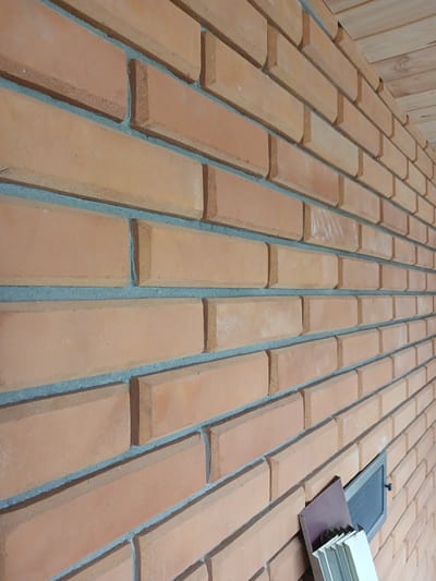 The image shows a close-up, angled view of an exterior brick wall. The bricks are a warm, reddish-orange color with visible gray mortar between them. The wall is constructed with a stretcher bond pattern. Towards the bottom right of the image, there are a few objects leaning against the wall: a dark reddish-brown rectangular object, and a stack of light-colored, multi-paneled objects. There is also a dark gray ventilation grate with a mesh screen embedded in the wall above these objects. The wall appears to be part of a building, and in the upper right corner, a portion of a wooden ceiling or soffit is visible, constructed from light-colored planks laid horizontally. The lighting suggests it is daytime, with shadows cast by the objects and the texture of the bricks. This image is described as being located in the city of Sharkawshchyna, Belarus.