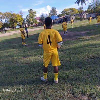The image shows a group of young men playing soccer in Nungwi, Tanzania. The coordinates provided locate the scene on a grassy field near the beach. The man in the center of the image, wearing a yellow jersey with the number 4, is standing on the field and looking towards the camera. Another man wearing a yellow jersey with the number 9 is walking away from the camera towards the right side of the image. Two additional men are visible in the distance wearing similar yellow jerseys. All men are wearing black shorts and white socks. The field is surrounded by trees and buildings. The sky is blue with a few white clouds. The image was taken with a Samsung Galaxy A05s phone.