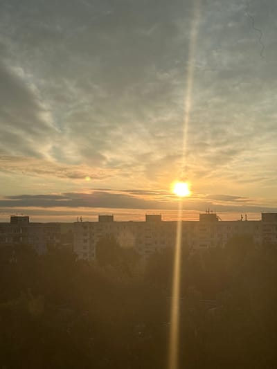 The image captures a sunrise over the city of Ozerny, Russia, with a bright sun positioned slightly left of center in the sky. The sun emits a strong beam of light that vertically bisects the frame, creating a lens flare effect. Scattered clouds with streaks of light fill the sky, suggesting a partly cloudy or hazy atmosphere. In the middle ground, apartment buildings with rows of windows are visible, interspersed with dense green trees. The foreground is dominated by dark foliage, likely trees or bushes, obscuring any ground-level details. The overall scene is bathed in the warm, golden light of dawn, casting a soft glow over the urban landscape. There are no discernible people or animals in the image.