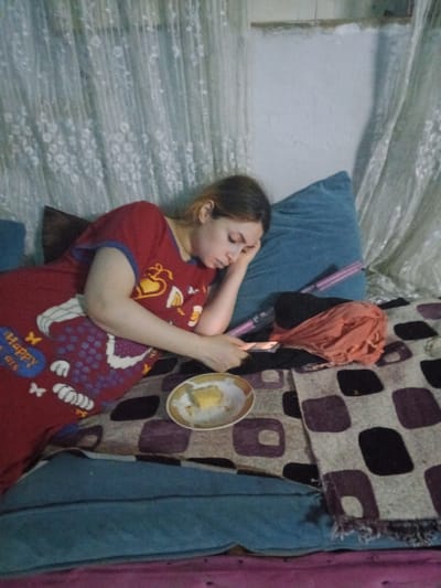 A young woman is lying on a couch, her head resting on a blue pillow. She is wearing a red and white patterned shirt. She is holding a black phone in her left hand, and her right hand is resting on the pillow. There is a plate with a piece of food in it in front of her. The couch is covered in a patterned blanket with a blue cushion underneath. There is a pink blanket on the couch.  The image is most likely taken in a home in Alexandria, Egypt (latitude: 31.227, longitude: 29.962).