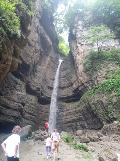 The image depicts a group of people at the Khushtyt waterfall in Khushto-Syrt, Russia. The waterfall cascades down a narrow, high canyon with layered rock walls covered in greenery. In the foreground, a person in a white t-shirt and dark pants is facing away from the camera. Further back, a person in a red t-shirt and dark pants stands near the base of the waterfall. To the right of the person in red, a woman in a light blue top, tan shorts, and a light yellow backpack is walking away from the camera, followed by a young girl in a pink t-shirt and purple leggings. The ground is a mix of dirt, rocks, and some vegetation. The overall environment is a natural, somewhat rugged setting with the dominant features being the waterfall and the canyon walls.