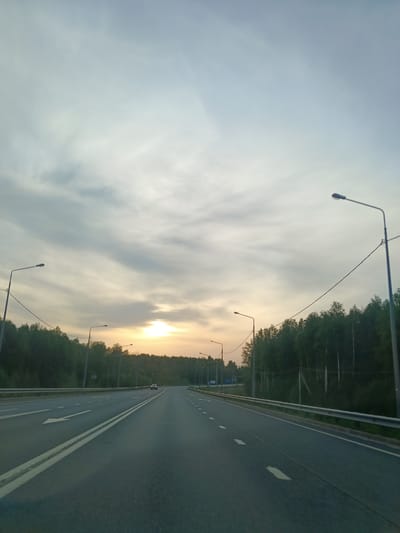 The image depicts a multi-lane highway in what appears to be a rural or suburban area of Mostovaya, Russia. The road is paved with asphalt and marked with white lines. A white arrow is painted on the leftmost lane, indicating a left turn. Street lamps line the sides of the road, and a metal guardrail is visible on the right. The edges of the highway are bordered by trees and vegetation. In the distance, a vehicle is driving on the road. The sky is cloudy with a hint of sunlight, suggesting it is either early morning or late evening. There are power lines running alongside the highway.