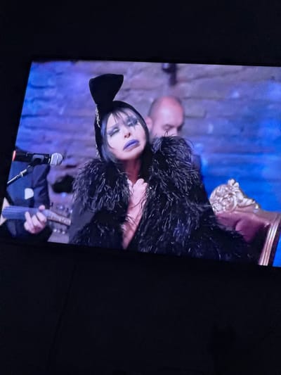 The image shows a woman with dramatic makeup, including purple lipstick and dark eye makeup, wearing a black outfit with feathered accents. She has a black headpiece with a bow. The woman is positioned in front of a microphone and appears to be holding a musical instrument, possibly a violin or similar stringed instrument. In the background, there is a man with a bald head and what seems to be an ornate chair. The setting appears to be indoors, possibly a stage or performance venue. The image is a photograph of a screen displaying this scene, with dark surroundings indicating the room where the photograph was taken. There is no mention of the city of Glojene, Bulgaria in the scene.