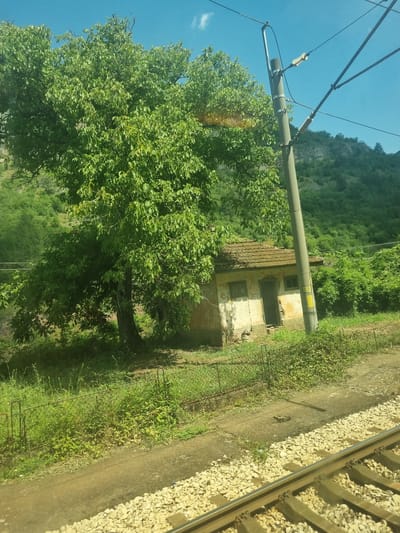 The image depicts an abandoned small, light-colored building with a tiled roof situated next to a large, leafy green tree. The building appears weathered and has two windows and a doorway. A utility pole with electrical wires stands to the right of the building and tree. The foreground shows railroad tracks and ballast. In the background, a lush, green hillside is visible under a bright blue sky with scattered clouds. The scene is set in a rural environment, potentially on the outskirts of Milanovo, Bulgaria, characterized by natural vegetation and infrastructure related to transportation. No people or animals are present in the image.