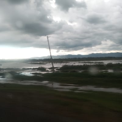 The image depicts an outdoor landscape, identified as Kobura ward, Kenya, under a heavily overcast sky. Dense, grey cloud cover dominates the upper portion of the frame, with a brighter, less opaque segment visible towards the upper left. A distant line of hills or mountains is silhouetted against the horizon. The midground features an extensive body of standing water, suggesting a flooded area or a large wetland. Partially submerged patches of green vegetation are distributed across this water. A single wooden utility pole, equipped with multiple overhead power lines, is positioned centrally in the midground, extending vertically from the water into the cloudy sky. Numerous small, dark, indistinct shapes are observable within the standing water in the central midground, suggesting a large group of birds or floating debris. The immediate foreground is dark and blurred, consistent with a road or elevated ground from which the photograph was taken, with faint light reflections in the lower right, possibly from water droplets on a window. No human subjects or distinct structures beyond the utility pole are visible.