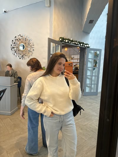 An indoor mirror selfie captures a woman in Kalinovo, Russia, standing in what appears to be a cafe or restaurant. The primary subject, a woman with long dark hair, is visible from the waist up, wearing a white ribbed sweater and light blue jeans. She holds an orange-cased smartphone with her right hand to take the photo, while her left hand is in her pocket. A black bag is slung over her left shoulder.

Reflected in the mirror behind her, another woman with brown hair, dressed in a light-colored sweater and blue jeans, is seen from behind, walking towards the left. Further left, a third woman in a dark jacket is seated at a light-colored reception desk. The background features light grey walls, a decorative starburst mirror, and a security camera mounted high on the wall. Two grey framed double doors, adorned with a white and silver festive garland, open to another section of the establishment where warm-toned hanging lights and blurred figures are visible. The flooring is light-colored, laid in a herringbone pattern.