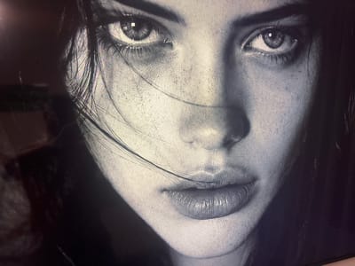 The image is a close-up, black and white portrait of a woman's face. The lighting is dramatic, with strong shadows and highlights that emphasize the contours of her features. Her eyes are open and looking directly at the camera, with intricate details visible in the irises and eyelashes. A few strands of dark hair fall across her face, partially obscuring her left eye. Her lips are full and parted slightly, with a glossy sheen that catches the light. The texture of her skin is highly detailed, with visible pores and subtle imperfections. The background is dark and indistinct, suggesting a studio setting or a dimly lit environment. There is no discernible location in the city of Krāslava, Latvia, visible or implied in this image.