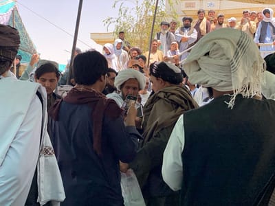 The image shows a crowd of men gathered in the city of Zaker-E Sharif, Afghanistan. The men are mostly wearing traditional Afghan clothing, including turbans and long shirts. Some men are wearing white turbans and are holding their hands together in a prayer position. One man in the foreground is taking a picture of another man with a smartphone. The scene appears to be taking place in a semi-open area. The background shows several men standing on a platform or a balcony. There are some buildings visible in the background as well. The overall mood of the scene is peaceful. The location indicated by the latitude and longitude coordinates (31.548, 65.76) corresponds to the city of Zaker-E Sharif, Afghanistan. The exact location within the city cannot be determined from the image.