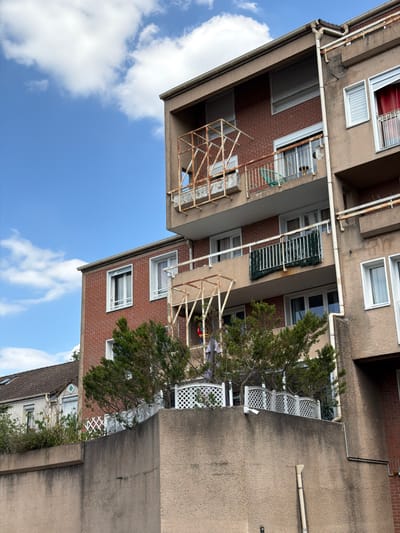 The image shows a residential building in Évry-Courcouronnes, France, on a clear day with scattered clouds. The building has a dated, Brutalist architectural style with a rough, textured brown facade and exposed brick sections. There are several balconies, some with rusty metal railings and one with a green fence-like covering. One balcony appears to be under renovation or in disrepair, with a scaffolding-like structure made of weathered metal. A red curtain is visible through a window on an upper floor. In the foreground, a concrete wall with some vegetation and a white lattice fence runs along the bottom of the frame. Adjacent to the main building, a smaller, pitched-roof house with a tiled roof is partially visible. The overall impression is of an older, potentially multi-unit residential area.