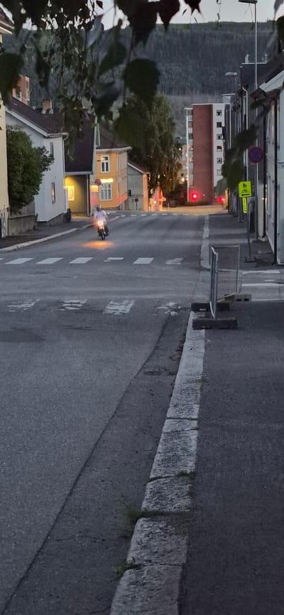 A motorcycle is riding down a street in Lillehammer, Norway. The scene is depicted at dusk, with streetlights and building lights illuminating the area. The road is paved and has white dashed lines marking the lanes. A stone curb separates the road from a sidewalk or embankment. On the left side of the road, there are several buildings, including a yellow building with orange trim and windows. A red traffic light is visible in the distance. A cyclist symbol and a "no stopping" sign are also present on the left side of the road. The motorcycle rider is wearing a white shirt and the motorcycle's headlights and taillights are on. Some foliage is visible in the foreground on the left side of the image. The weather appears to be calm. No specific emotions are conveyed in the image.