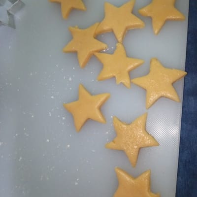 The image shows a collection of star-shaped cookies made from dough, arranged on a white cutting board. Some of the cookies are slightly imperfect, with some edges appearing less defined. A silver star-shaped cookie cutter is partially visible in the upper left corner. The background consists of a textured white surface, likely the cutting board, with small white specks scattered across it, possibly flour. To the right of the cutting board, a dark blue textured surface, possibly fabric, is visible. The lighting is somewhat dim, casting subtle shadows on the cookies and the cutting board. There are no people or explicit indications of the location within the city of El Limón, Venezuela, evident in the image itself. The scene appears to be a close-up, overhead view of food preparation.
