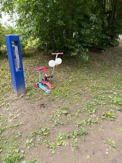 The image captures an outdoor scene in Tokkari-Lend, Russia, under natural daylight, likely on an overcast day or with dappled light. The ground is a blend of brown dirt and patchy green grass, dotted with various weeds and a few small yellow dandelions. Dense green foliage from bushes and trees forms a lush backdrop along the left side of the frame.

Three children's scooters are present in the mid-ground, appearing to be casually left on the ground. One scooter is pink with white handles. Another, positioned behind it, is red and black. To its right, a blue and pink scooter with a cartoon character design on its deck rests on the grass.

In the foreground, towards the bottom left, there is a blue rectangular structure. It appears to be a freestanding unit, possibly a small bench or a dispenser, with an open section containing several white papers or leaflets, though the text on them is not legible. No people are visible in the scene. The setting suggests a casual children's play area, possibly in a park or a communal garden, currently quiet and unoccupied.