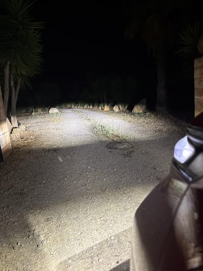The image depicts a gravel road at night, illuminated by the headlights of a vehicle that is partially visible on the right side of the frame. The road is lined with rocks and sparse vegetation, with palm trees visible in the background. The scene appears to be a rural or semi-rural area, consistent with the location of Santa Margalida, Spain. There are no people visible in the image. The overall impression is one of a quiet, dimly lit, and undeveloped area.