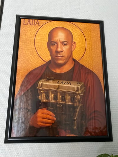The image shows a framed portrait of a bald, muscular man resembling Vin Diesel, depicted in an artistic style reminiscent of religious icons. He has a halo of gold mosaic behind his head and wears a dark garment draped over his shoulders. In his hands, he holds a silver-colored car engine part, possibly a cylinder head, with the word "LADA" inscribed on it. The word "LADA" is also written in red text at the top left of the image, above the man's head. The background is a golden mosaic pattern. The frame is black. The photograph is taken indoors, with a textured white wall visible in the background outside the frame. There is no discernible information within the image or crops to suggest a specific location in Seleevo, Russia.