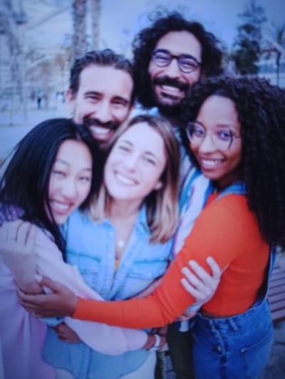 In a close-up, low-resolution image, five individuals are captured in a group embrace, their faces turned towards the camera and smiling. The group consists of three women and two men. The individuals are diverse in terms of apparent ethnicity and gender. They appear to be outdoors, with blurred foliage and palm trees visible in the background, suggesting a park or an outdoor recreational area. The lighting appears to be natural daylight. The setting does not provide any specific visual cues that would indicate the location as Balakliya, Ukraine. The people are dressed casually in various colored shirts and denim overalls. The interaction is one of friendship and closeness.