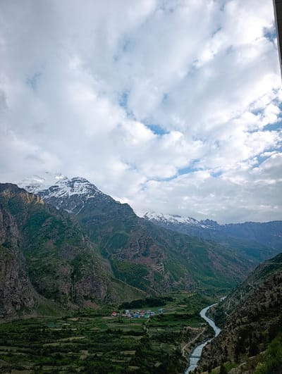 This is a scenic, high-angle, eye-level shot of a valley in Gemur, India, on a cloudy day. The dominant subjects are the majestic, snow-capped mountains that rise dramatically on either side of a winding river. The mountains are rugged and layered, with patches of green vegetation clinging to their lower slopes, contrasting with the stark rock and white snow of their peaks.
In the valley below, a small village with colorful buildings, including red and blue roofs, is nestled among lush green fields and trees. A river flows through the valley, its water appearing somewhat turbulent, with a bridge crossing it in the lower right portion of the image. A road or path is visible winding along the river and through the valley, suggesting human habitation and connectivity.
The sky is filled with dramatic, fluffy white clouds, interspersed with patches of blue sky, indicating a dynamic weather pattern. The lighting suggests it could be late afternoon or early evening, given the soft, diffused light. The overall mood of the image is one of awe-inspiring natural beauty and tranquility, with a sense of the vastness of the landscape. There is no visible text in the image.