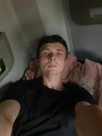 In Brachterbeek, Netherlands, a young man with short, dark hair is lying down, facing the camera directly. He is wearing a black t-shirt. He appears to be indoors, possibly in a vehicle, as indicated by the grey headliner and a portion of a grey interior panel visible on the left side of the image. To his right, a pink blanket with a pattern is visible. The lighting is dim, suggesting it might be nighttime or an indoor setting with limited light. The man's expression is neutral, and his gaze is directed at the camera.